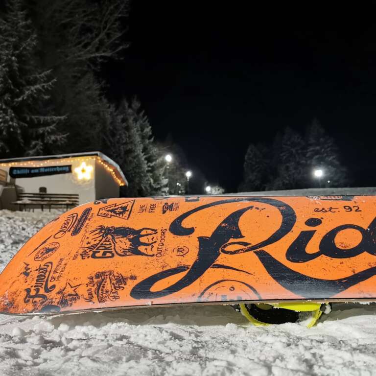 Snowboard im Schnee vor der beleuchteten Piste