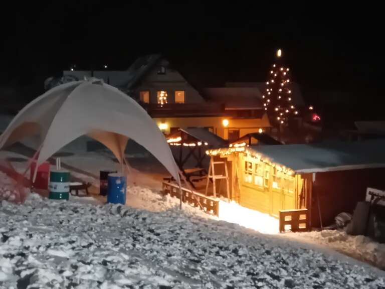 Die beleuchtete Glühweinalm am Abend, daneben ein geschmückter Weihnachtsbaum
